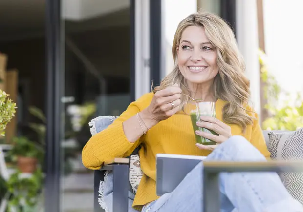 A smiling woman wearing a yellow sweater sits comfortably in a chair, holding a glass of green drink and a spoon. She appears relaxed, enjoying her beverage in a cozy setting with plants in the background.