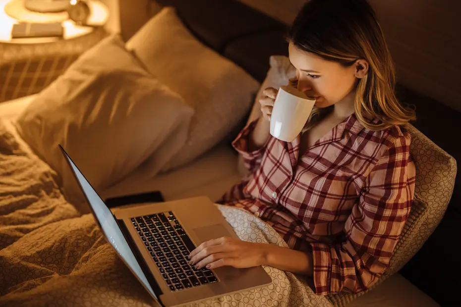 Жена пие топла напитка и чете на лаптоп