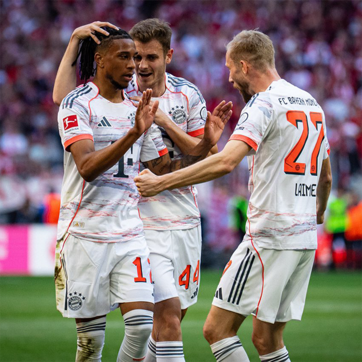 Футболистите на Bayern Munich празнуват след гол. Трима играчи с бели и оранжеви екипи се радват, като един от тях е с дредове. Атмосферата е енергична и емоционална, показвайки радост и отборен дух.