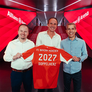 Трима мъже позират пред червена стена с надпис "Allianz Arena". Един от тях държи футболнa фланелка на FC Bayern München с надпис "2027 DOPPELHERZ". Всички изглеждат усмихнати и доволни.