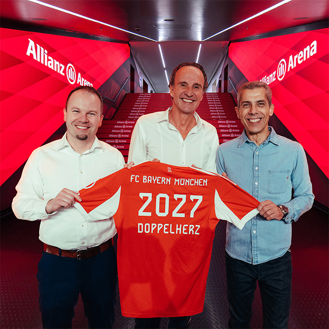 Трима мъже позират пред червена стена с надпис "Allianz Arena". Един от тях държи футболнa фланелка на FC Bayern München с надпис "2027 DOPPELHERZ". Всички изглеждат усмихнати и доволни.