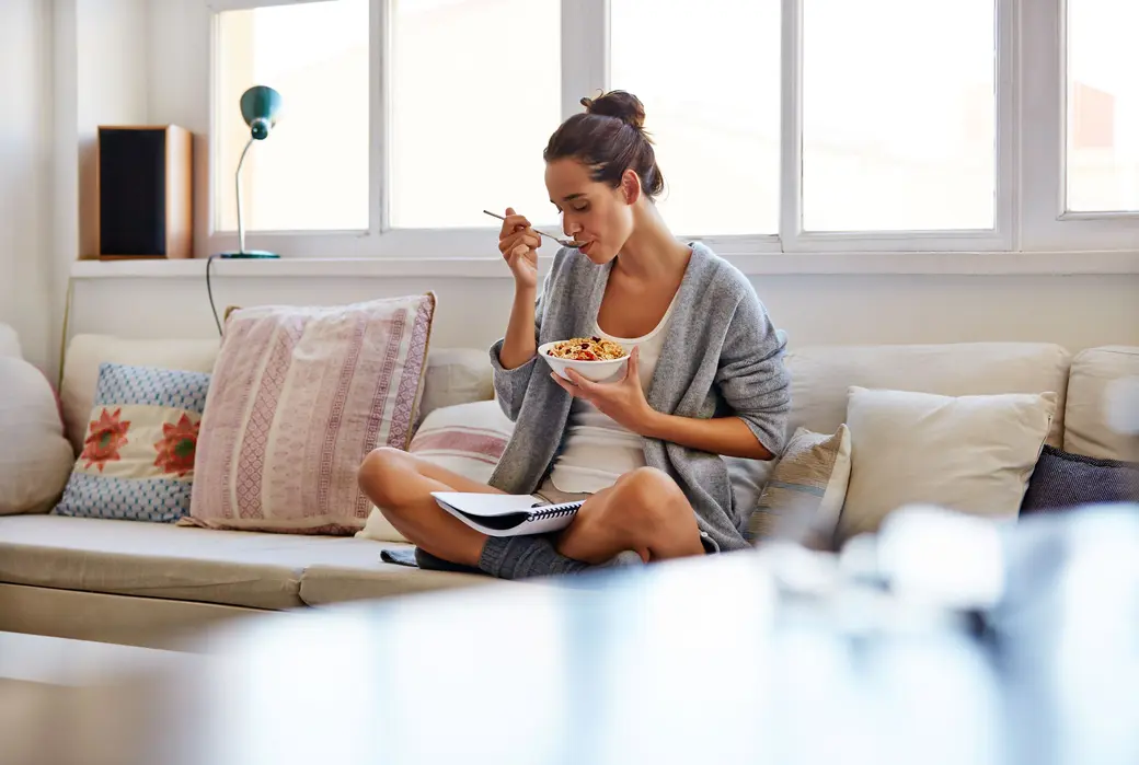 Жена седи на диван и яде йогурт, докато чете