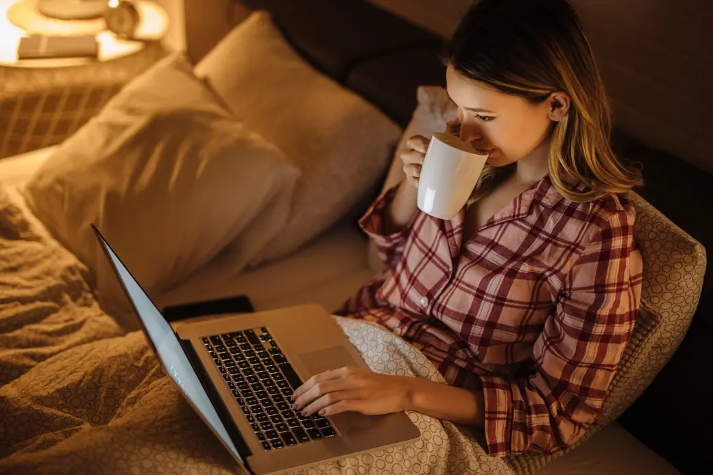 Жена с пижама работи на лаптоп в леглото, докато пие топла напитка